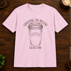 Surviving the Present T-Shirt in Light Pink on a dark wood coffee bar