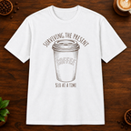Surviving the Present T-Shirt in White on a dark wood coffee bar