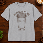 Surviving the Present T-Shirt in Sport Grey on a dark wood coffee bar